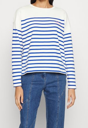 Osoba ubrana w biały top z długim rękawem z poziomymi niebieskimi paskami oraz niebieskie dżinsy, stojąca ze zrelaksowanymi ramionami wzdłuż ciała.