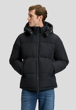 Mann mit schwarzer Steppjacke mit Kapuze, Hände in den Taschen, blickt nach links, trägt blaue Jeans vor hellem, schlichtem Hintergrund.