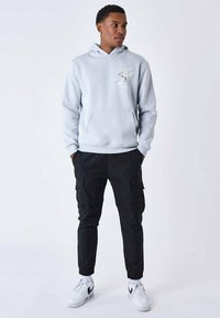 Sweatshirt à capuche gris clair avec poche kangourou et détail graphique, associé à un pantalon cargo noir et des baskets blanches. Coupe décontractée, design minimal.