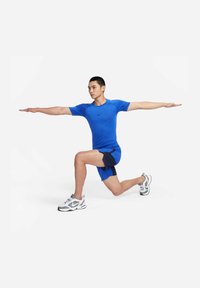 Blå idretts skjorte med korte ermer og en tettsittende design, kombinert med marinebl�å shorts. Modellen står i en lungeposisjon og har på seg løpesko.