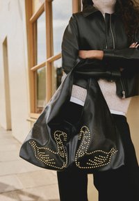 Zwarte leren slonzige tas met een gouden zwanenontwerp met studs. Heeft een brede opening en een zachte textuur, gedragen door een persoon in een zwart jack.