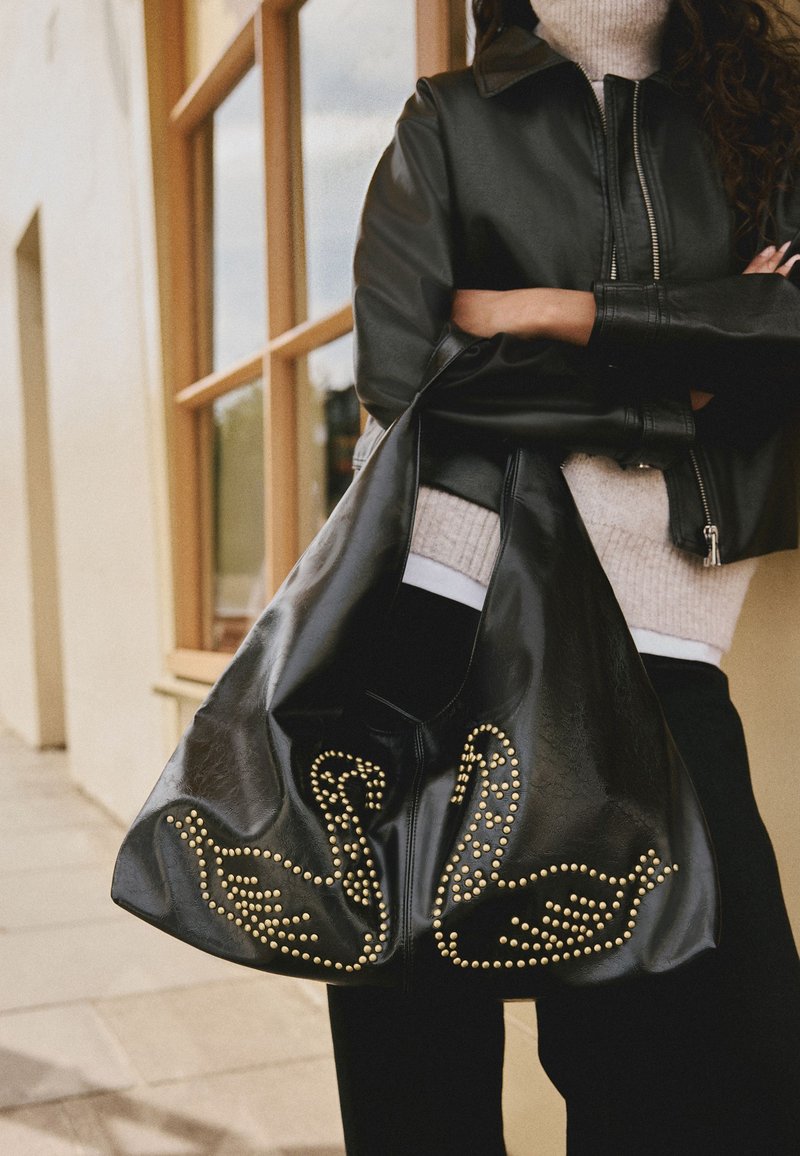 Zwarte leren slonzige tas met een gouden zwanenontwerp met studs. Heeft een brede opening en een zachte textuur, gedragen door een persoon in een zwart jack.