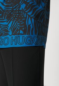 Korte mouwen shirt met een blauw tropisch bladerenpatroon op zwarte stof, met een gedurfde "HUGO" logo aan de onderrand. Matt textuur, relaxte pasvorm.