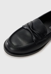Mocassino in pelle nera con dettaglio della fascia annodata, superficie liscia, punta arrotondata e soletta imbottita, con una suola in gomma a profilo basso.
