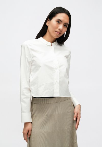 Λευκό κοντό πουκάμισο με μακριά μανίκια και όρθιο γιακά. Λείο ύφασμα με διακριτική λάμψη. Συνδυάζεται με μπεζ φούστα ριμπ.