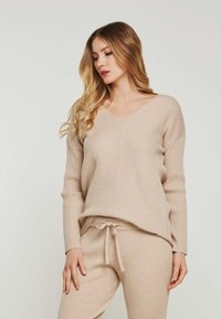 Beige gerippter Strickpullover mit V-Ausschnitt und langen Ärmeln, kombiniert mit passenden Jogginghosen mit Kordelzug. Die strukturierte Stoffqualität verleiht Volumen.