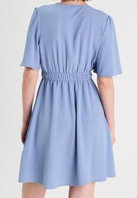 Lysblå kjole lavet af blødt stof. Har korte puffærmer, en samlet talje og en udsvingende nederdel, der falder over knæet.