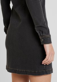 Musta denimi mekko, jossa on pitkät hihat, istuva malli ja napitettu hihansuu. Huomattava kontrastistiikka helman ja hihojen varrella.