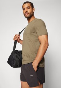 T-shirt ajustado verde oliva e calções pretos com um logótipo. O modelo carrega uma mala de desporto preta com um painel lateral texturizado.