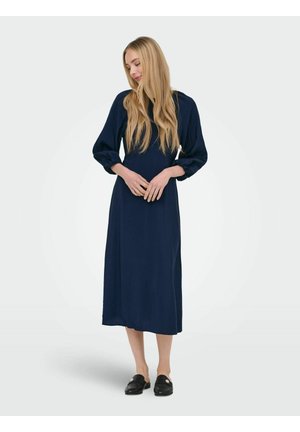 Femme aux cheveux blonds longs portant une robe midi bleu marine à manches bouffantes et des chaussures noires faciles à enfiler, debout les mains jointes.