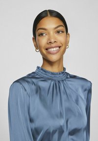 Donna con occhi marroni e capelli scuri raccolti in uno chignon basso, che indossa orecchini a cerchio d'oro e una blusa di raso blu con collo alto, che sorride leggermente guardando a destra.