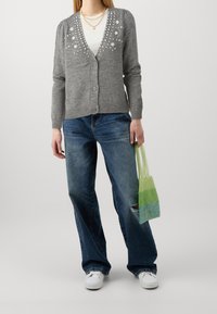 Kobieta w szarym swetrze zapinanym na guziki z perłowymi koralikami, białej bluzce, rozerwanych szerokich dżinsach i białych trampkach, trzymająca małą zieloną torebkę z koralików.