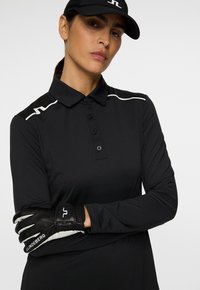 Eine Frau trägt ein schwarzes Langarm-Polo, eine schwarze Kappe und einen schwarzen Golfhandschuh mit weißen Logos, während sie ihre Arme vor einem schlichten Hintergrund verschränkt.