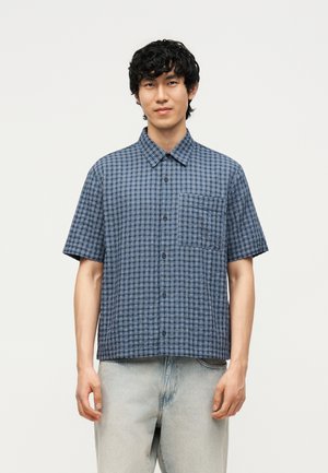 Młody mężczyzna z kręconymi czarnymi włosami, ubrany w niebieską kraciastą koszulę z krótkim rękawem i jasne dżinsy na jednolitym tle.