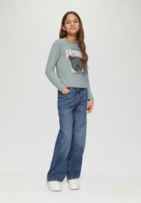 Lange mouwen lichtgrijs shirt met zwart luipaard grafiek en roze tekst, gecombineerd met losse blauwe denim jeans en witte sneakers.