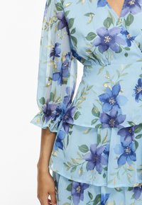 Blouse bleu clair avec manches longues transparentes et imprimé floral violet avec feuilles vertes et accents dorés, portée sur le haut du corps d'une personne.