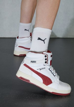 Person trägt weiße und rote Puma Slipstream High-Top-Sneaker mit weißen Puma-Logo-Socken auf dunklem Boden.