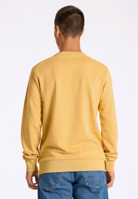 Gul sweatshirt med långa ärmar, ribbad halsringning och muddar, med en klassisk passform. Kombinerad med blå denimjeans, som visar en enkel baksidedesign.