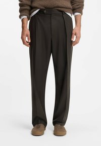 Pantaloni tagliati di colore marrone scuro con una parte frontale piegata, realizzati in un tessuto liscio. Presentano una chiusura con un bottone singolo e gamba affusolata, abbinati a scarpe chiare.