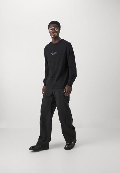 Sudadera negra con un logo azul, combinada con pantalones cargo negros. El modelo lleva zapatos negros brillantes. Fondo liso.