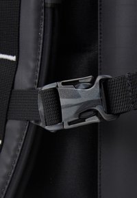 Czarny materiałowy pasek plecaka z szarym plastikowym zapięciem w kamuflażu, z regulowanym splotem i teksturowanym wykończeniem.