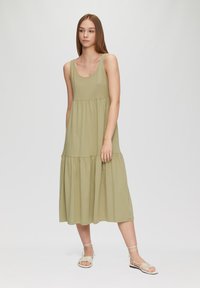 Robe midi vert olive avec un décolleté arrondi, jupe à volants et texture douce. Associée à des sandales à brides de couleur claire.