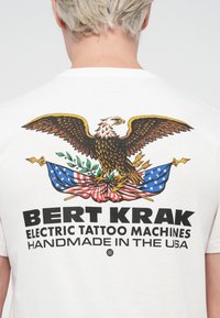 White cotton t-shirt featuring a colorful eagle design, U.S. flag accents, and bold black text reading "BERT KRAK Electric Tattoo Machines."