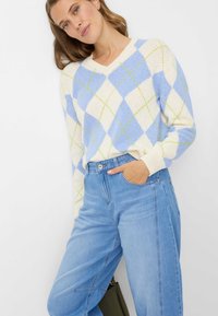 V-hals, gebreide trui in lichtblauw en crème, met een ruitpatroon met groene accenten. Gecombineerd met een high-waisted, lichtgewassen spijkerbroek.