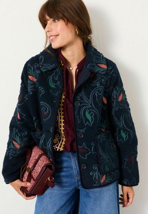 Navy fleece jacket with floral embroidery in green, red, and orange. Features a collar, buttons, and two front pockets. Worn over a burgundy top.