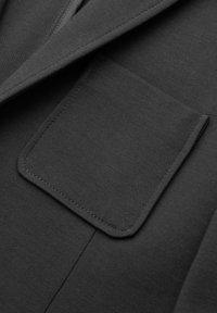 Poche en tissu gris foncé avec un rabat assorti sur un vêtement gris foncé, montrant une couture fine et un matériau texturé.