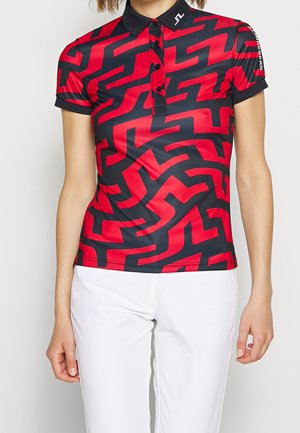 Vrouw draagt een rood-zwart poloshirt met korte mouwen en een abstract doolhofachtig patroon, gecombineerd met een witte broek, staand tegen een effen achtergrond.