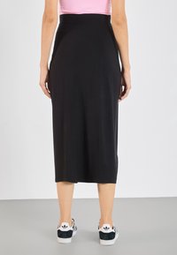 Frau trägt einen schwarzen Midirock, ein rosa Oberteil und weiße Adidas-Sneaker, steht mit dem Rücken zur Kamera auf einem grauen Boden vor einer weißen Wand.