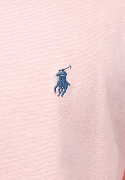Μπλε κεντημένο λογότυπο ενός παίκτη πόλο πάνω σε άλογο με υψωμένη ρακέτα σε απαλό ροζ ύφασμα.