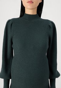 Pull en tricot côtelé vert foncé avec un col haut, des manches bouffantes et un tissu texturé. Gros plan mettant en évidence le motif et la forme du tricot.