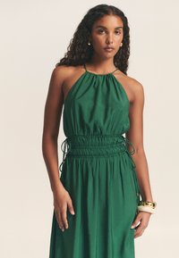 Zelené šaty na zavazování za krk se sbíraným výstřihem a stahovacím páskem v pase. Vyrobené z hladké tkaniny, s plynulou siluetou a minimálními detaily.