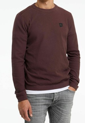 Homme portant un pull marron foncé texturé à manches longues sur une chemise blanche avec un jean gris clair, main dans la poche, sur fond blanc.