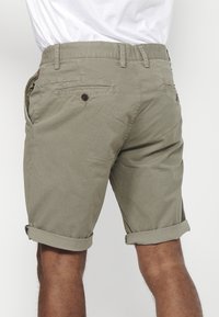 Man in opgerolde beige shorts en een wit t-shirt, gezien van achteren op dijhoogte tegen een effen achtergrond.