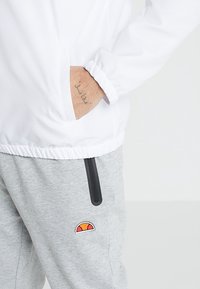 White, lightweight jacket with elastic cuffs and waistband, paired with gray sweatpants featuring a small, colorful logo patch and side zipper pockets.
