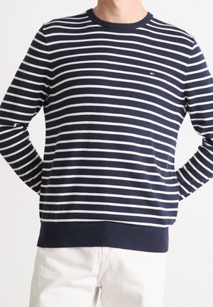 Homme portant un pull à rayures horizontales bleu marine et blanc à manches longues avec un petit logo sur la poitrine et un pantalon blanc sur fond uni.