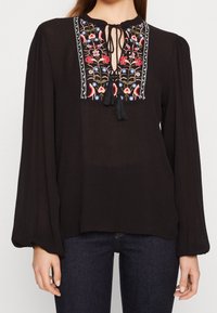 Svart blus med långa, voluminösa ärmar. Har en nyckelhålsutskärning med färgglad blommönsterbroderi och fransdetaljer längs öppningen.