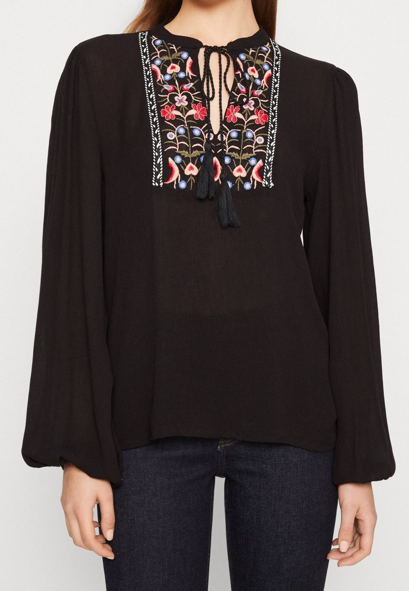 Svart blus med långa, voluminösa ärmar. Har en nyckelhålsutskärning med färgglad blommönsterbroderi och fransdetaljer längs öppningen.
