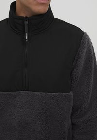 Sort og grå fleecepullover med høj krave, der har lynlåslukning og en tekstureret, fluffy nederste sektion. Med logo-drag.