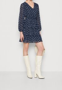 Modré šaty s vzorem námořnického stylu, dlouhými rukávy, výstřihem do V a vrstevnatým lemem; spárované s bílými kozačkami po kolena a malou bílou kabelkou.