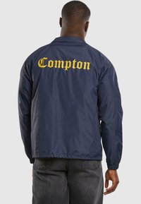 Jachetă de tip windbreaker de culoare bleumarin, cu text galben "Compton" pe spate, manșete elastice și tiv drept. Material neted și ușor.