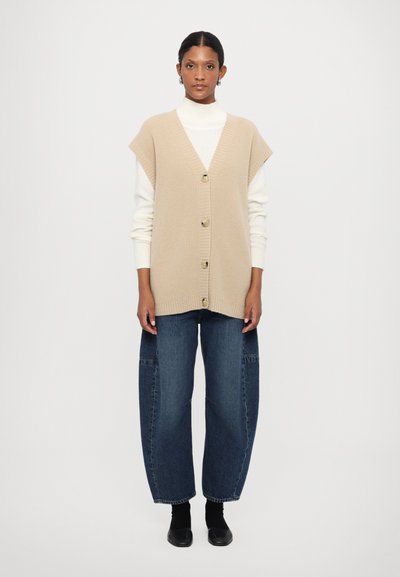 Beige ribbet cardigan vest med V-hals, forsynet med fire store knapper og korte ærmer, kombineret med løse blå denimbukser.