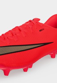 Φωτεινό κόκκινο ποδοσφαιρικό παπούτσι Nike με μαύρο λογότυπο swoosh, κορδόνια και διαμορφωμένες τάπες στη σόλα, εμφανιζόμενο σε γωνιακή κοντινή λήψη.