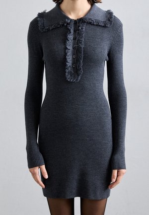 Femme portant une robe ajustée en maille côtelée gris foncé, manches longues, col à franges et patte boutonnée sur le devant.