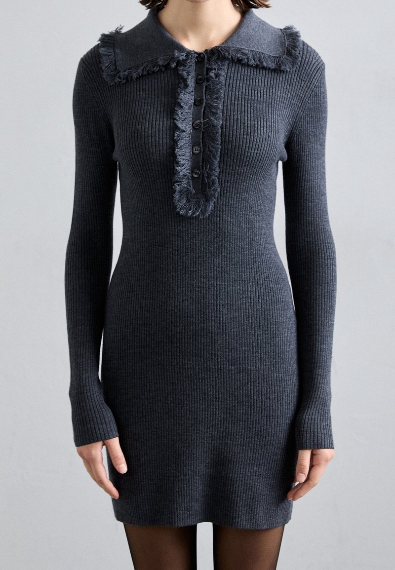 Femme portant une robe ajustée en maille côtelée gris foncé, manches longues, col à franges et patte boutonnée sur le devant.