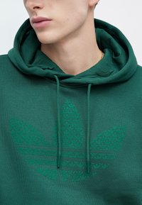 Πράσινο φούτερ με υφασμένη, τονική μοτίβα λογότυπου. Διαθέτει κουκούλα με κορδόνι και απαλό ύφασμα, κατάλληλο για καθημερινή χρήση.