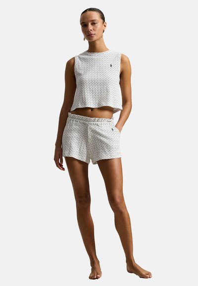 Femme debout pieds nus portant un haut court sans manches blanc et un short assorti avec un petit motif floral sombre sur un fond uni.